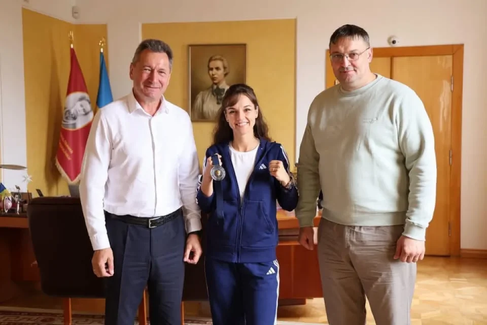 Жінка в синьому спортивному костюмі тримає медаль між двома чоловіками в кабінеті