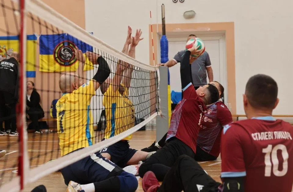 Гравці у бордових і жовтих футболках грають у волейбол сидячи в залі
